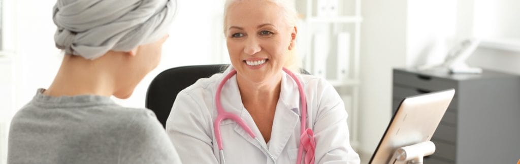 Two women are sitting in an office. The woman on the left is wearing a grey top and has a turban wrapped around her head. Her hands are being held by a smiling woman who is wearing a white lab coat and a pink stethoscope. Terri Crudup and a team of researchers have demonstrated the importance of a holistic approach to cancer treatments, otherwise known as integrative oncology, reviewing the efficacy of complementary therapies in breast cancer patients.